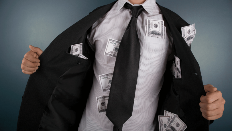 Um homem de negócios vestido com roupas formais está no foco da imagem. Ele abre seu terno e mostra várias notas de dólares espalhadas pelos seus bolsos.