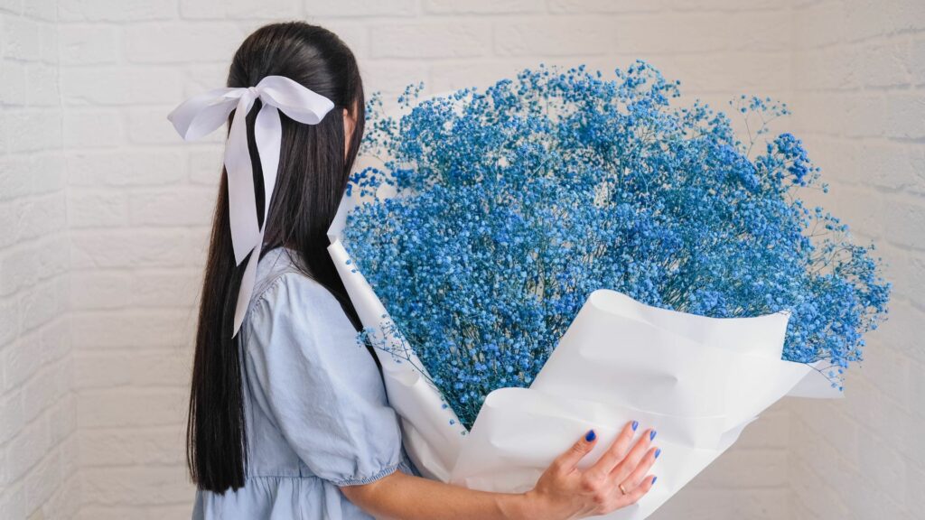 Imagem de uma mulher  com um laço de fita branco na cabela, segurando um buquê de Gypsophila ou "véu de noiva" na cor azul.
