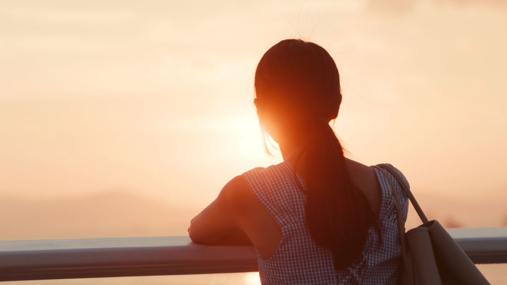 Uma mulher está de costas para a imagem, porém está de frente para o pôr do sol. Ela observa-o e está encostada em uma grade.