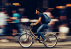 Um homem está andando de bicicleta em uma rua movimentada e está escutando música nos seus fones de ouvido. Há pessoas caminhando atrás dele.