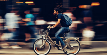 Um homem está andando de bicicleta em uma rua movimentada e está escutando música nos seus fones de ouvido. Há pessoas caminhando atrás dele.
