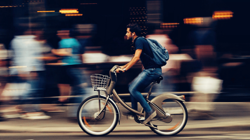 Um homem está andando de bicicleta em uma rua movimentada e está escutando música nos seus fones de ouvido. Há pessoas caminhando atrás dele.