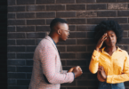 Um casal, composto por uma mulher e um homem, estão tendo um conversa séria. A mulher coloca a mão na cabeça, indicando estar incomodada, enquanto o homem fala.