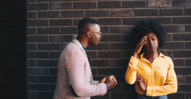 Um casal, composto por uma mulher e um homem, estão tendo um conversa séria. A mulher coloca a mão na cabeça, indicando estar incomodada, enquanto o homem fala.