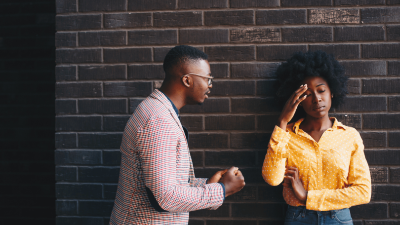 Um casal, composto por uma mulher e um homem, estão tendo um conversa séria. A mulher coloca a mão na cabeça, indicando estar incomodada, enquanto o homem fala.