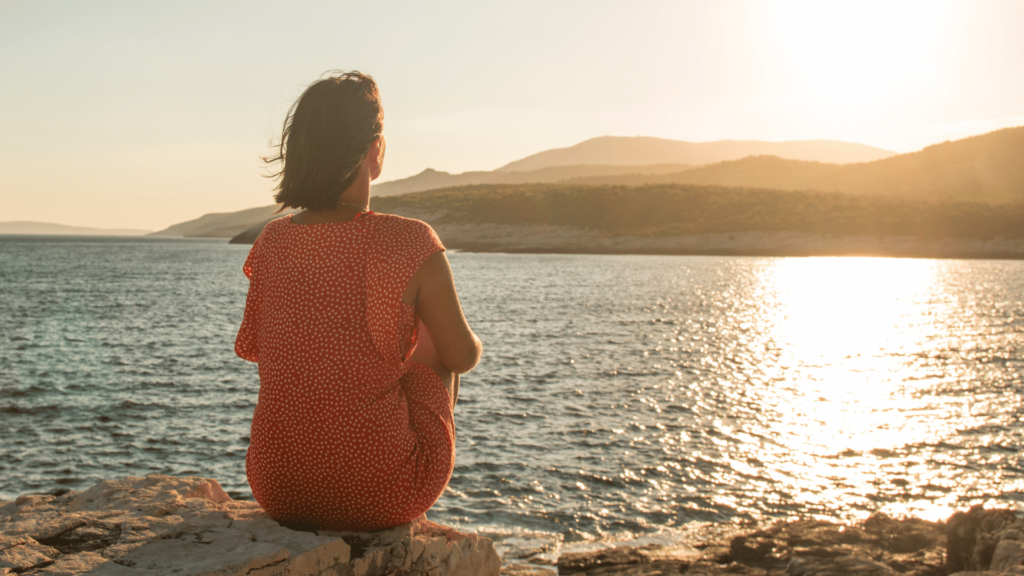 Uma mulher está sentada em uma pedra de frente para o mar e olhando reflexiva para o pôr do sol.
