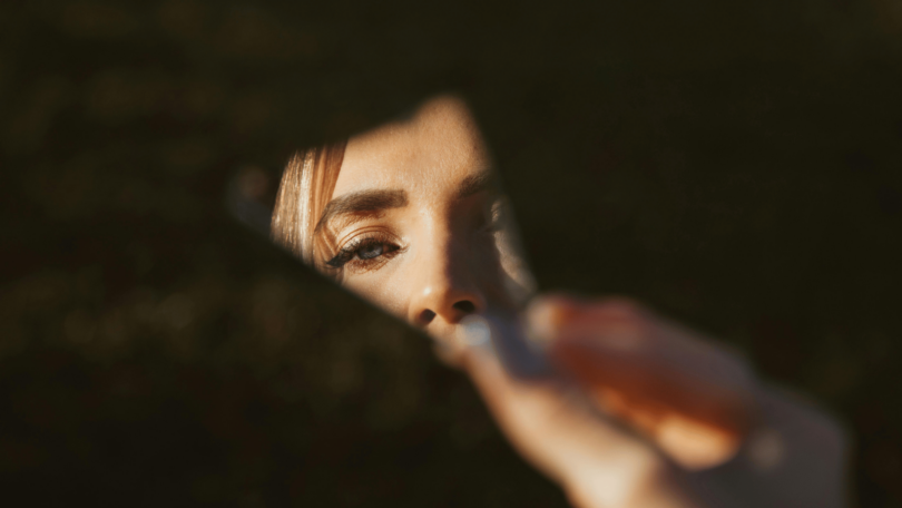 Uma mulher segura um fragmento de um espelho, o qual reflete uma parte do seu rosto.
