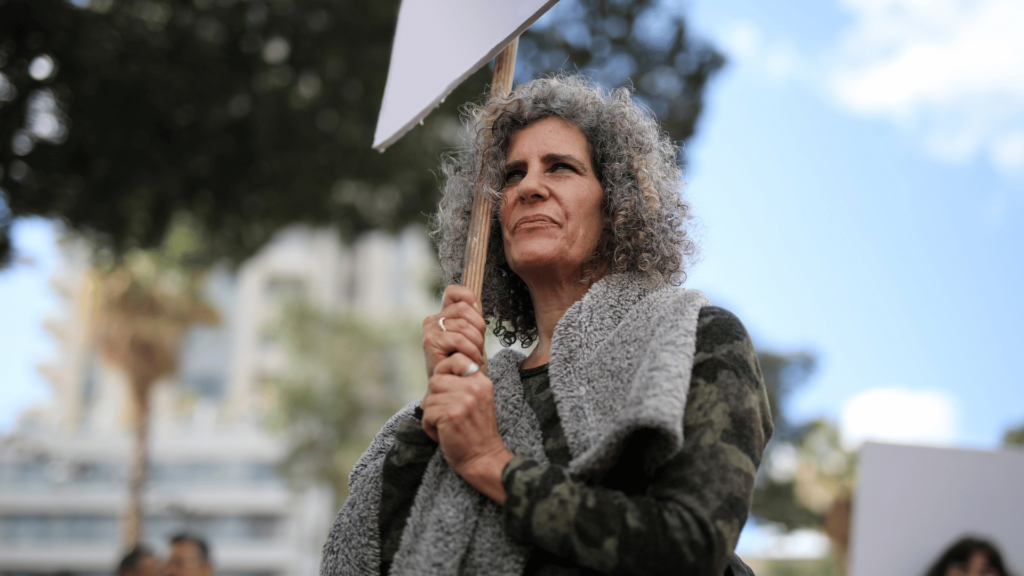 Uma mulher mais madura está protestando em uma rua com um cartaz na mão.