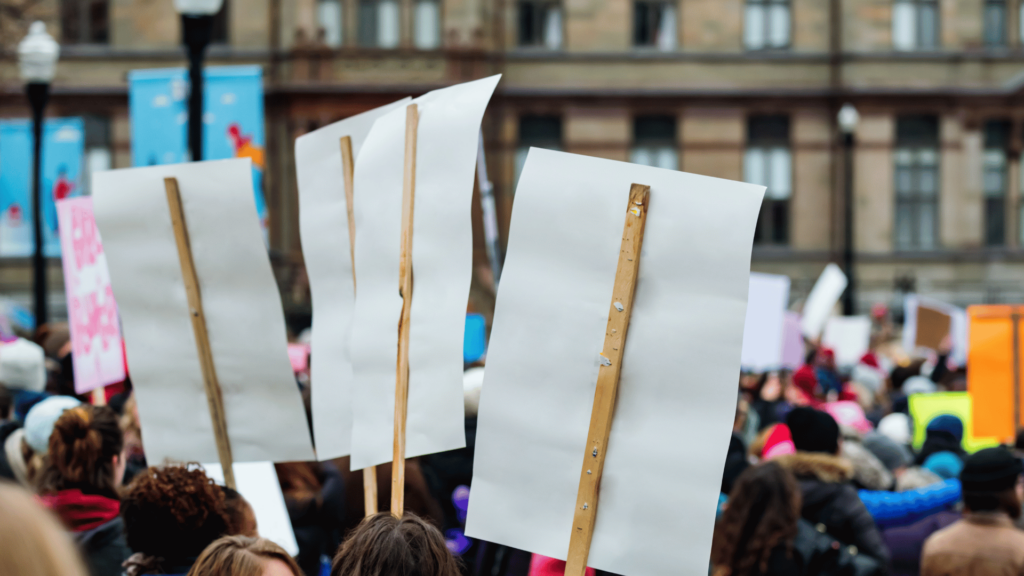 Um grupo grande de pessoas está se manifestando em uma cidade. Há cartazes sendo levantados por elas.