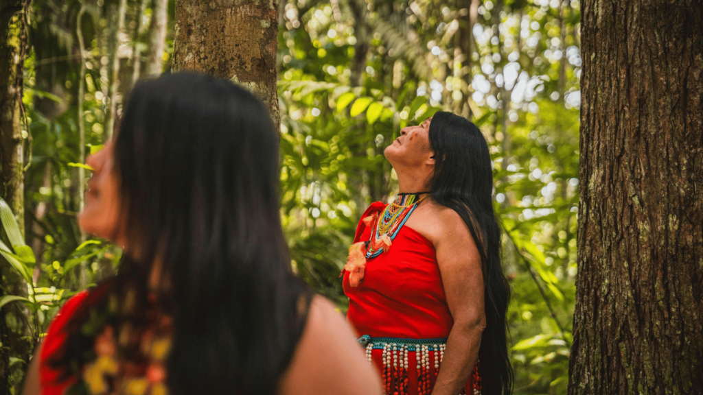 Duas mulheres indígenas estão no foco da imagem. Elas andam dentro da floresta e olham para cima.