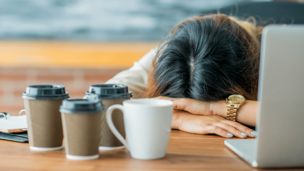 Uma mulher está curvada sobre uma mesa de trabalho, indicando estar cansada. Em cima da mesa, há três copos e uma xícara de café.