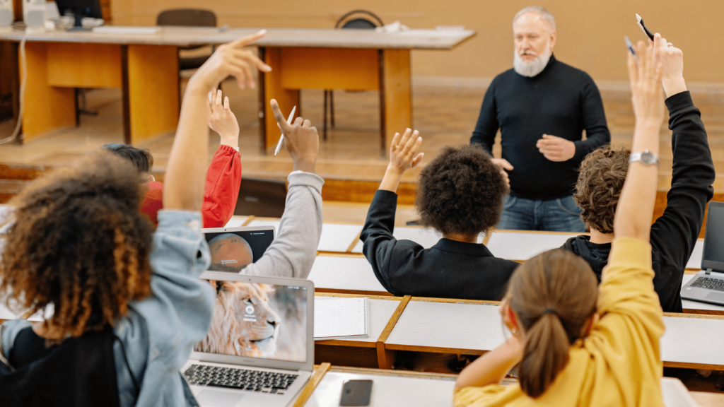 Na imagem, há vários alunos levantando suas mãos para falar em uma sala de aula. Na frente deles, o professor fala e expõe a aula.