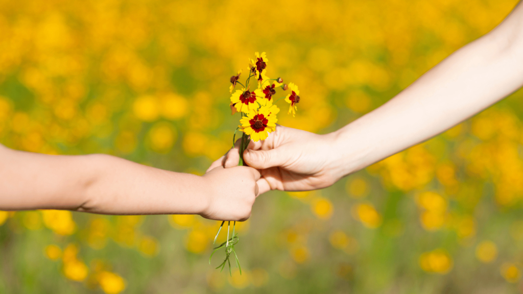 Uma criança entrega um conjunto de flores amarelas para sua mãe. Na imagem, apenas os braços dos indivíduos e as flores estão em destaque.