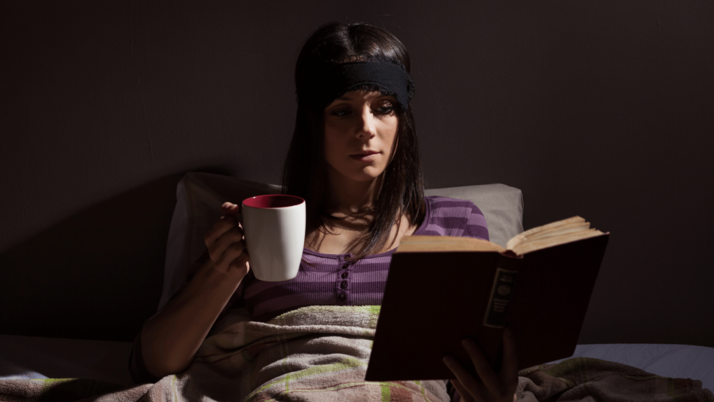 Uma mulher busca relaxar antes de dormir. Ela está sentada na sua cama lendo um livro e tomando algo em uma caneca.