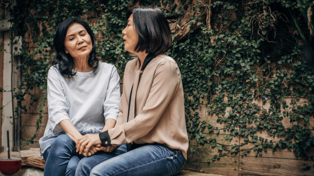 Duas mulheres asiáticas estão sentadas em um banco conversando. Uma segura a mão da outra.