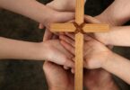Imagem de 4 pessoas, de mãos para cima, segurando uma cruz de madeira, simbolizando a fé e a liberdade.