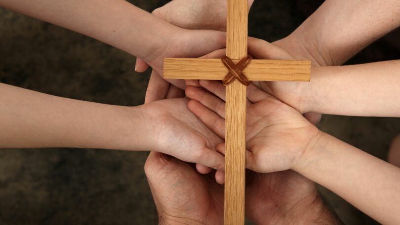 Imagem de 4 pessoas, de mãos para cima, segurando uma cruz de madeira, simbolizando a fé e a liberdade.