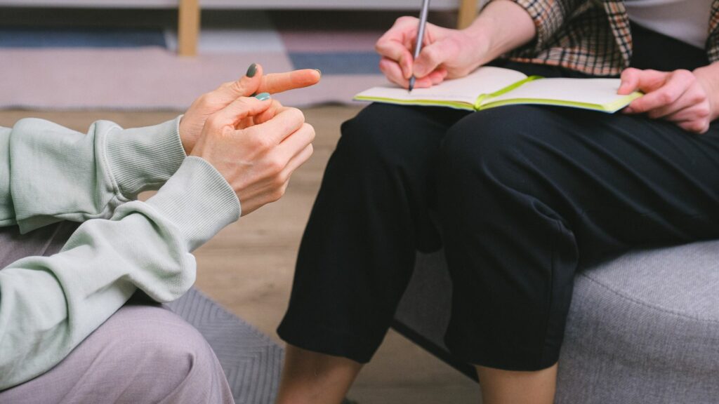 Imagem de duas mulheres conversando em uma sessão de terapia, sendo uma a paciente e a outra a terapeuta que está fazendo anotações em seu caderno.
