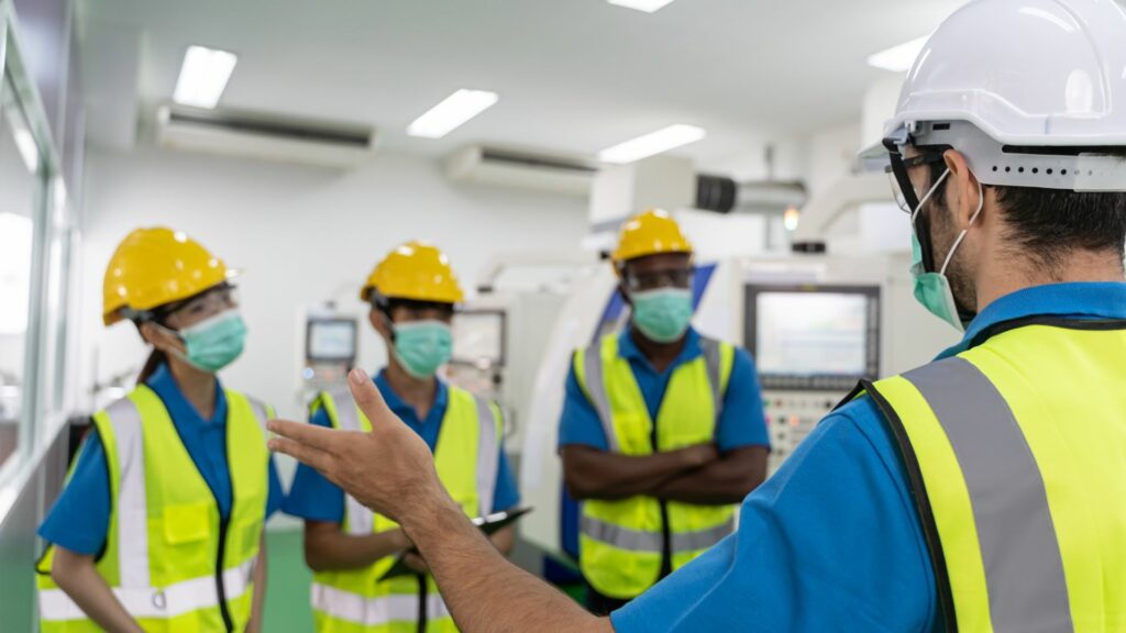 Imagem de um gestor e seus funcionários dentro de uma sala na empresa. Eles estão usando equipamentos de segurança e conversando. A foto traz o conceito de segurança e saúde no trabalho.