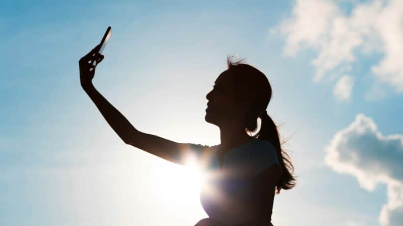 Imagem de fundo das nuvens no céu azul. Em destaque uma mulher fazendo uma selfie para postar nas redes sociais, trazendo o conceito de validação, viralização.