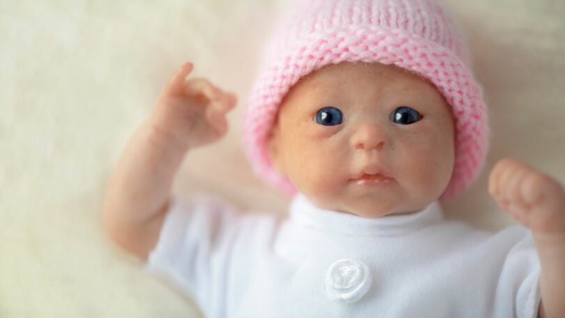 Imagem de um bebê reborn com uma camiseta branca e uma touca de tricot na cor rosa.