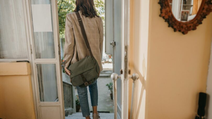 Imagem de uma mulher na fase adulta saindo da casa dos pais. Ela está de costas, com uma bolsa pendurada nos ombros.