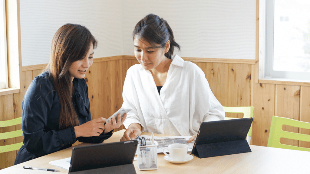 Duas mulheres asiáticas e de negócios estão trabalhando juntas. Há dois "tablets", além de canetas, papéis e uma xícara em cima de uma mesa.