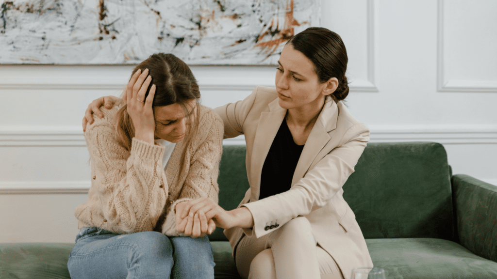 Duas mulheres estão sentadas em um sofá. Uma delas está cabisbaixa e triste. A outra mulher está apoiando-a e consolando-a.