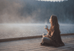 Uma mulher está relaxando em um ambiente de natureza. Ela está sentada, tomando um café e observando o pôr do sol.