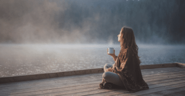Uma mulher está relaxando em um ambiente de natureza. Ela está sentada, tomando um café e observando o pôr do sol.