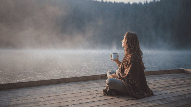 Uma mulher está relaxando em um ambiente de natureza. Ela está sentada, tomando um café e observando o pôr do sol.