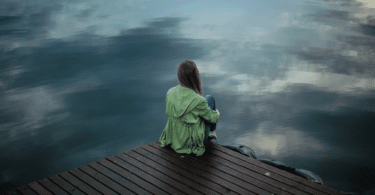Uma mulher jovem está sentada sobre uma ponte de madeira. Ela está contemplando o rio e a natureza ao seu redor.