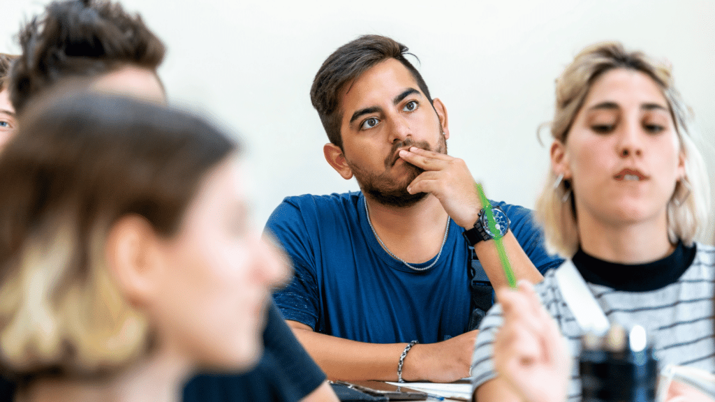Um estudante está prestando a atenção na aula. Ao redor dele, há outros estudantes. Todos estão em uma sala de aula.
