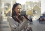 Uma mulher jovem está falando ou gritando em um megafone. Ela está na calçada de uma rua.