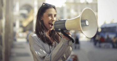 Uma mulher jovem está falando ou gritando em um megafone. Ela está na calçada de uma rua.