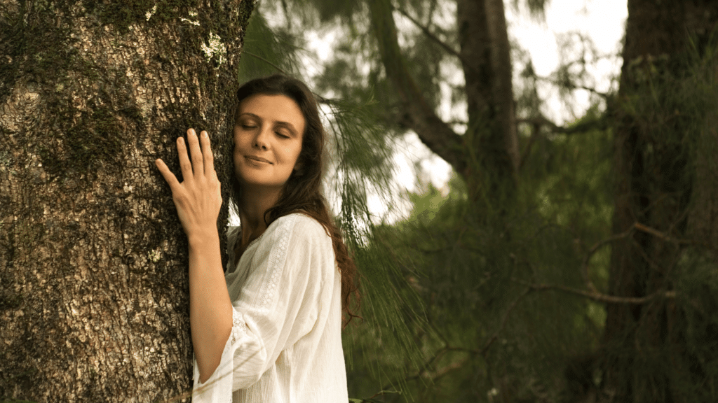Uma mulher jovem está com os olhos fechados e abraçando uma árvore. Ela está em um ambiente de natureza.