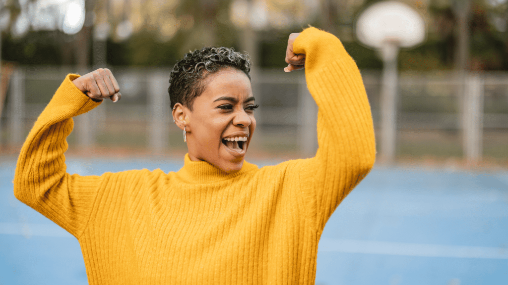 Uma mulher jovem, negra e de cabelos curtos está com os dois braços levantados simbolizando força.