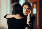 Duas amigas estão se abraçando. Uma está de costas para a imagem. A outra mulher, que está de frente para a imagem, tem a expressão que aparenta estar com intenções ruins com a amiga ou está sendo falsa.