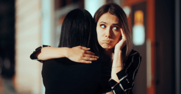 Duas amigas estão se abraçando. Uma está de costas para a imagem. A outra mulher, que está de frente para a imagem, tem a expressão que aparenta estar com intenções ruins com a amiga ou está sendo falsa.