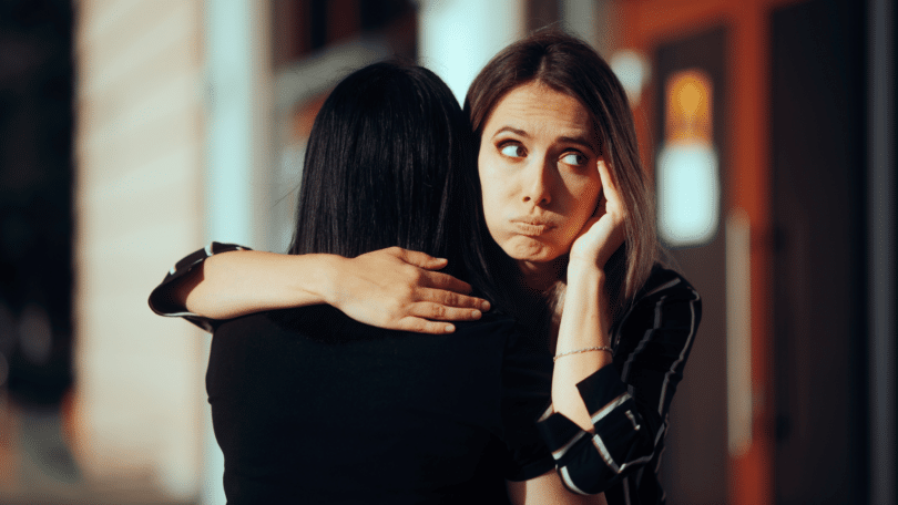 Duas amigas estão se abraçando. Uma está de costas para a imagem. A outra mulher, que está de frente para a imagem, tem a expressão que aparenta estar com intenções ruins com a amiga ou está sendo falsa.