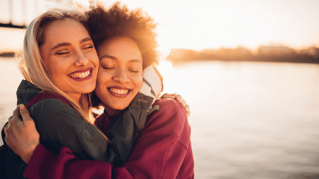 Duas amigas estão se abraçando e sorrindo. Elas estão em um ambiente de natureza. Atrás delas, há um rio e reflete a luz do pôr do sol.