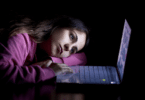 Uma mulher jovem está cabisbaixa e triste olhando e mexendo no seu notebook.