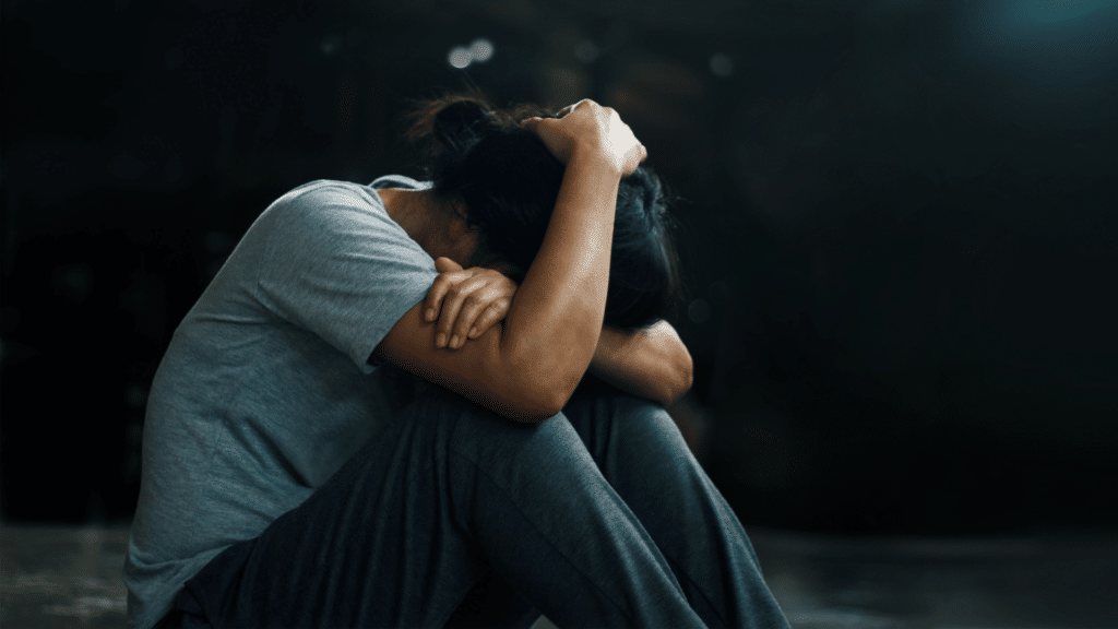 Uma mulher está encolhida e sentada no chão em um ambiente aberto, indicando que ela está triste, sofrendo ou deprimida.