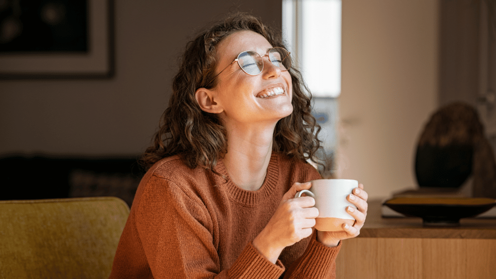 Uma mulher jovem está sentada na sua sala tomando café e sorrindo.
