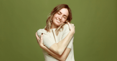 Uma mulher jovem está se abraçando e sorrindo, indicando autocuidado.