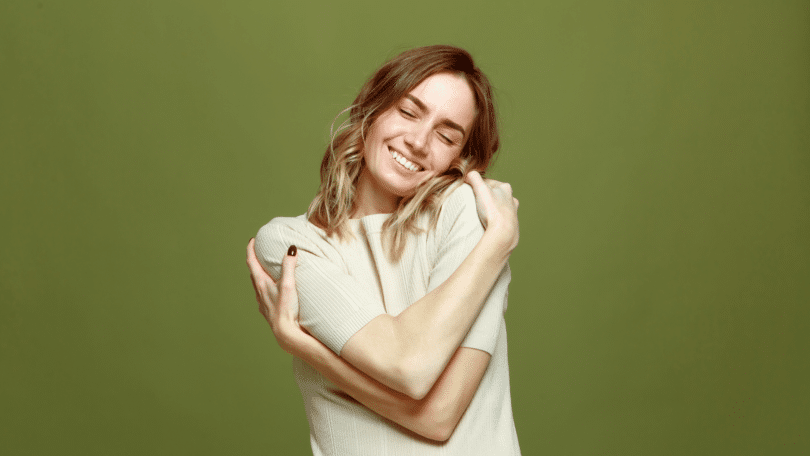 Uma mulher jovem está se abraçando e sorrindo, indicando autocuidado.