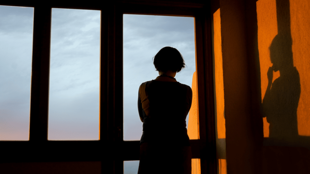 Uma mulher está de frente para uma janela. Ela olha pela janela, contempla a vista e está pensativa.