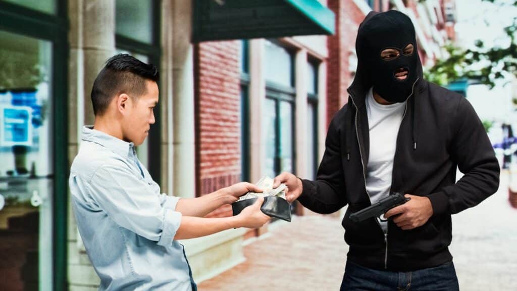Imagem de um homem assaltando um outro homem. Ele está roubando a carteira com dinheiro e aponta uma arma para a vítima. O assalto ocorre em uma rua.