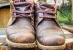 Imagem de um sapato velho, uma bota marrom, sobre um latão de ferro. O sapato está com marcas e sujo de barro.