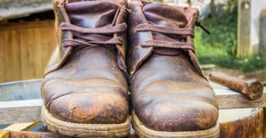 Imagem de um sapato velho, uma bota marrom, sobre um latão de ferro. O sapato está com marcas e sujo de barro.